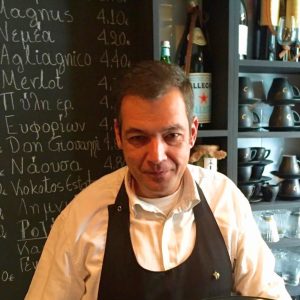 Cinque Wine & Deli team member in front of Greek wine list chalkboard inside wine bar in Psyrri Athens.