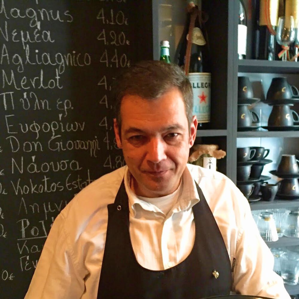 Cinque Wine & Deli team member in front of Greek wine list chalkboard inside wine bar in Psyrri Athens.