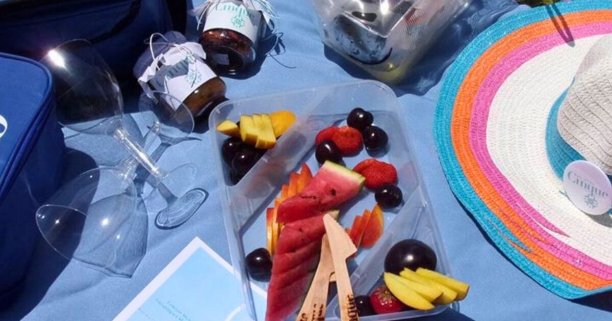 Cinque Mykonos tailor-made wine picnic basket with Greek wine bottle, fresh fruit platter, wine glasses and branded accessories set up outdoors on grass.