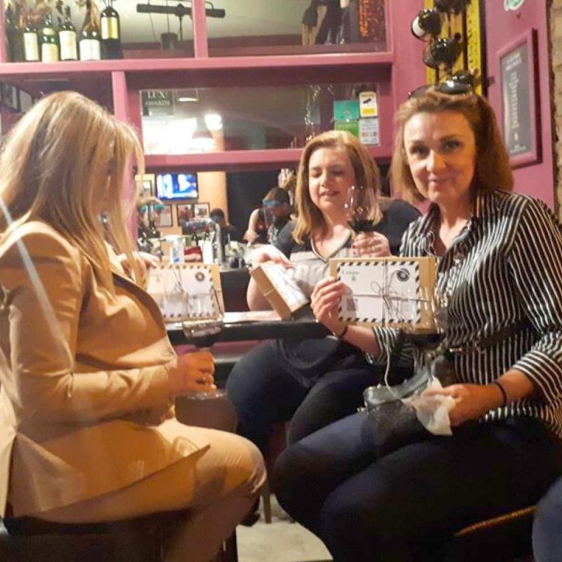 Guests enjoying red wine during an intimate tasting event at Cinque Wine & Deli in Athens, holding wine glasses and tasting boxes in a cozy wine bar setting.