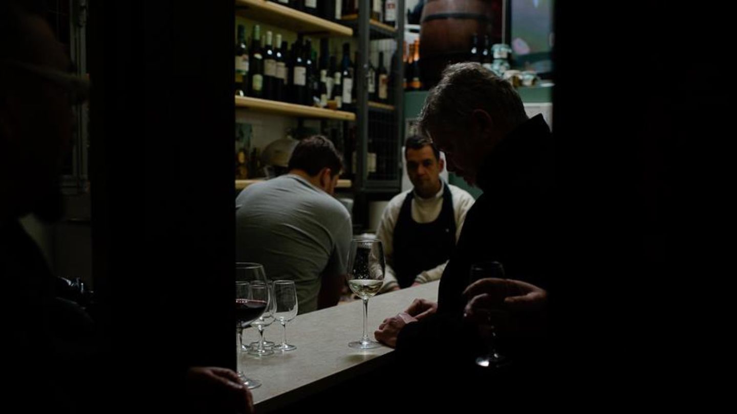 Atmospheric scene inside a small wine bar with guests tasting wine and shelves filled with Greek wine bottles in the background.