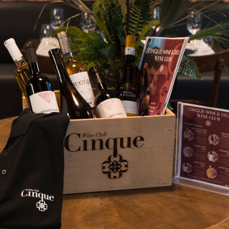 Cinque wooden box filled with Greek wines, Wine Club materials, and branded apparel, displayed on a table with deli and product development items.
