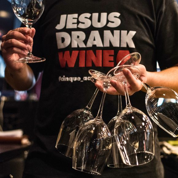 Person holding wine glasses wearing a Jesus Drank Wine t-shirt