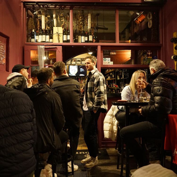 Guests enjoying wine at a cozy, bustling Cinque wine bar with warm lighting, lively conversations, and shelves filled with Greek wines.