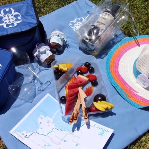 Cinque Mykonos tailor-made wine picnic basket with Greek wine bottle, fresh fruit platter, wine glasses and branded accessories set up outdoors on grass.