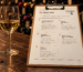 The Cinque Cellar wine list on a wooden table beside a glass of wine, in an illustrative cellar setting.